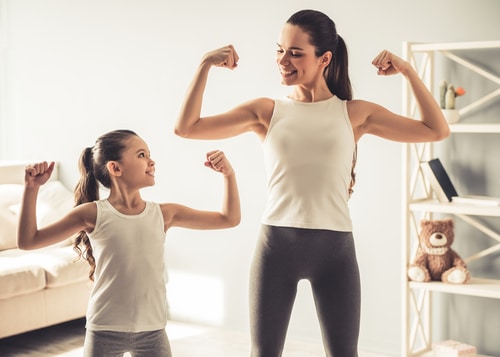 young woman and charming little daughter are showing their biceps and smiling while working out at home-img-blog