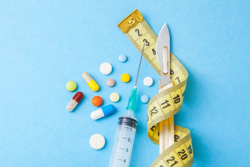 Collection of pain medication next to a syringe, scalpel, and tape measure against blue background.