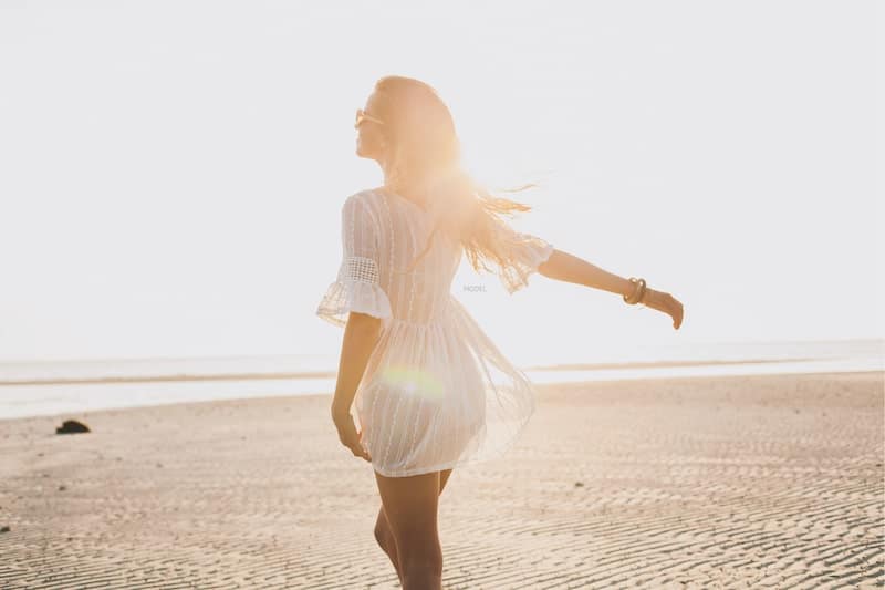 Silhouette of the woman's back side standing on a sunny beach wearing a thin white sun dress.