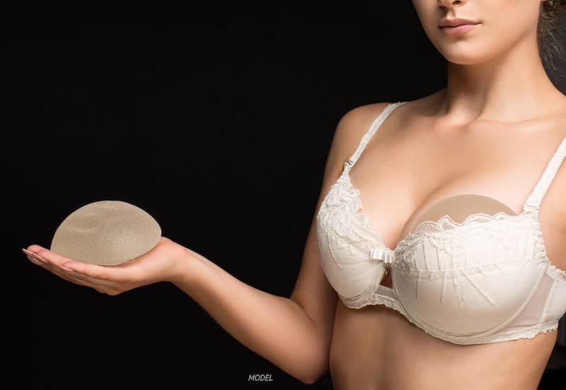 A woman wearing a bra and holding a breast implant in her hand.