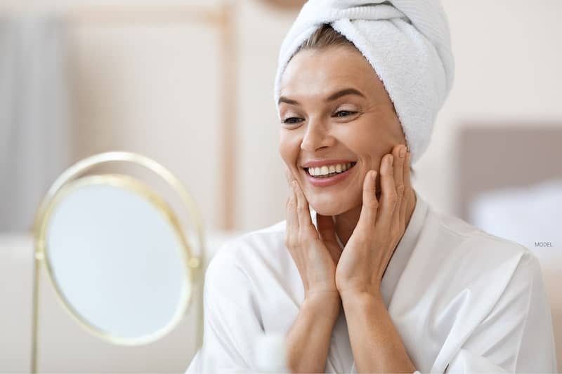Beautiful middle-aged woman in a bathrobe with a towel on her head smiling at her reflection in the mirror